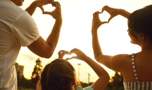 A family of 3 holding up hand shaped hearts to the sky